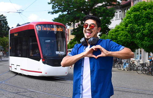 Junger Mann mit Sonnenbrille steht vor einer Straßenbahn und formt mit den Händen ein Herz.